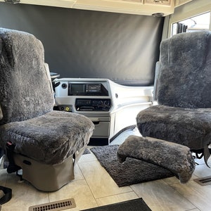 May include: Two gray faux fur seat covers for a vehicle. The covers are installed on the front seats of a motorhome. The seats are facing each other.