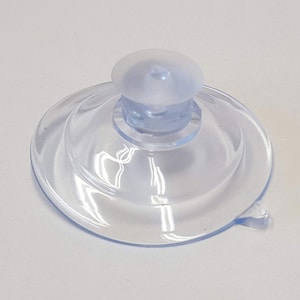 May include: A clear, transparent suction cup with a rounded top and a flat, circular base. The suction cup is made of a flexible material and has a small tab on the side for easy removal. The design is simple and functional.