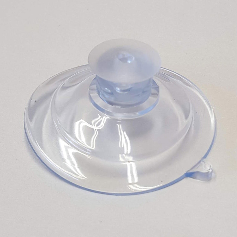 May include: A clear, transparent suction cup with a rounded top and a flat, circular base. The suction cup is made of a flexible material and has a small tab on the side for easy removal. The design is simple and functional.