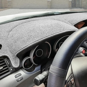 May include: A gray dashboard cover is installed in a car. The cover is made of a soft, felt-like material and fits over the dashboard. The steering wheel is black leather with a perforated design. The car's dashboard gauges are visible.