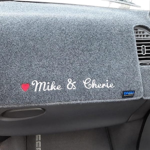 May include: A gray dashboard cover with the embroidered names "Mike & Cherie" and a red heart. The cover is designed to fit a car's dashboard and has a vent opening.