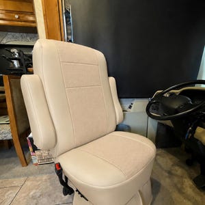 May include: A beige captain's chair with a smooth leather-like finish on the sides and a textured fabric center panel. The seat features integrated armrests, a high backrest, and a skirted base, positioned next to a black steering wheel.
