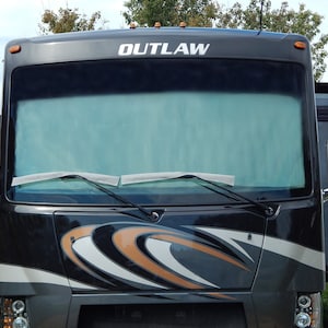 May include: Front view of a black and gray Outlaw RV. The windshield wipers are visible, and the word "OUTLAW" is printed in white across the top. The RV has orange and white decorative accents.