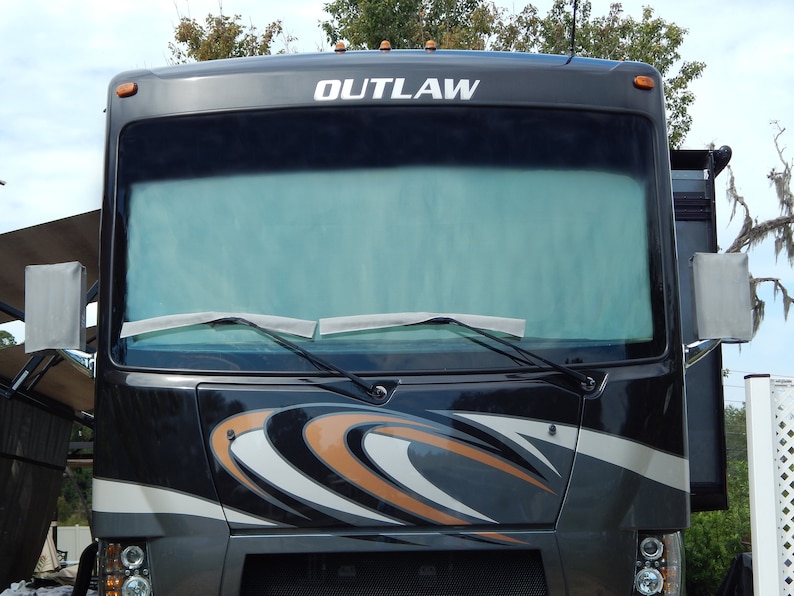 May include: Front view of a black and gray Outlaw RV. The windshield wipers are visible, and the word "OUTLAW" is printed in white across the top. The RV has orange and white decorative accents.