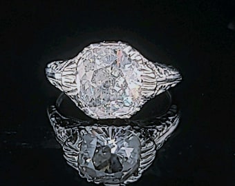 Könnte beinhalten: Nahaufnahme eines Diamantrings mit aufwendiger Filigranfassung. Der große, klare Diamant ist der Mittelpunkt und reflektiert das Licht. Der Ring ist vor einem dunklen Hintergrund platziert, was seine Brillanz hervorhebt.
