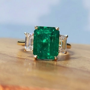 Op de afbeelding: Een gouden ring met een grote smaragd geslepen groene edelsteen en twee kleinere smaragd geslepen heldere edelstenen aan weerszijden.