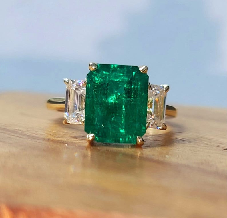 Op de afbeelding: Een gouden ring met een grote smaragd geslepen groene edelsteen en twee kleinere smaragd geslepen heldere edelstenen aan weerszijden.