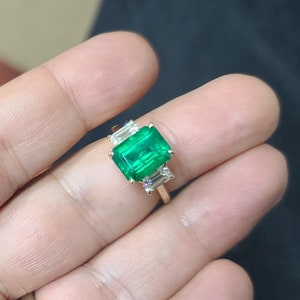 Op de afbeelding: Een gouden ring met een grote smaragdgroene edelsteen omringd door drie kleinere, heldere edelstenen.