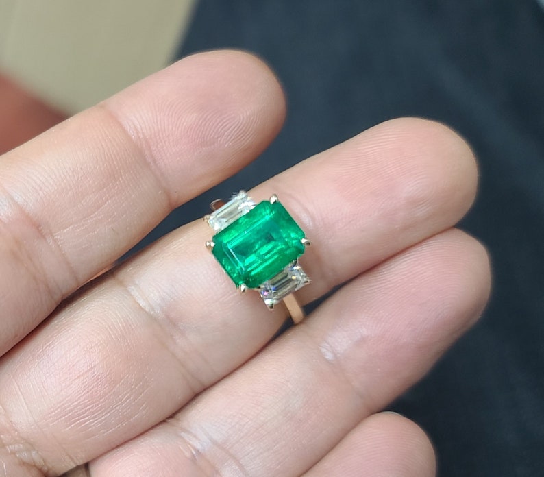 Op de afbeelding: Een gouden ring met een grote smaragdgroene edelsteen omringd door drie kleinere, heldere edelstenen.