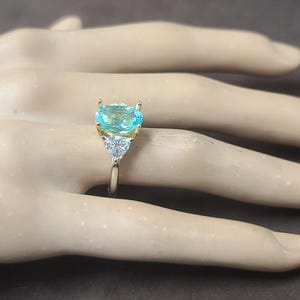 May include: A gold ring featuring a large, light blue gemstone with a rectangular cut. Two smaller, triangular white gemstones flank the main stone. The ring is set on a gold band, creating a striking contrast.