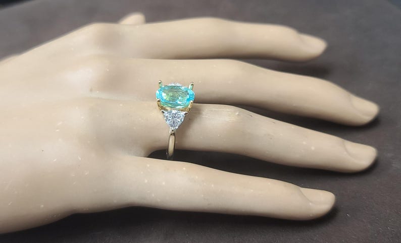 May include: A gold ring featuring a large, light blue gemstone with a rectangular cut. Two smaller, triangular white gemstones flank the main stone. The ring is set on a gold band, creating a striking contrast.