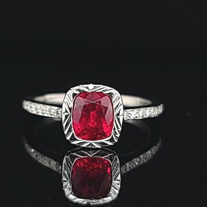 May include: A silver ring with a red gemstone set in a square-shaped bezel. The ring is accented with small diamonds around the band.
