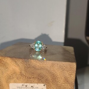 May include: A silver ring with a large, oval-cut, light blue gemstone as the centerpiece. Two smaller, triangular-shaped, clear gemstones flank the main stone. The ring is set on a wooden block, showcasing its design and sparkle.