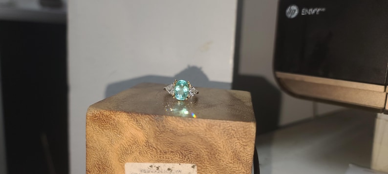 May include: A silver ring with a large, oval-cut, light blue gemstone as the centerpiece. Two smaller, triangular-shaped, clear gemstones flank the main stone. The ring is set on a wooden block, showcasing its design and sparkle.