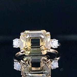 May include: A gold ring with a large, light yellow, emerald-cut gemstone surrounded by two smaller, round, clear gemstones.