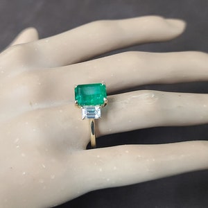 Op de afbeelding: Een gouden ring met een grote smaragdgroene edelsteen en twee kleinere, heldere edelstenen. De ring is op een hand van een mannequin.