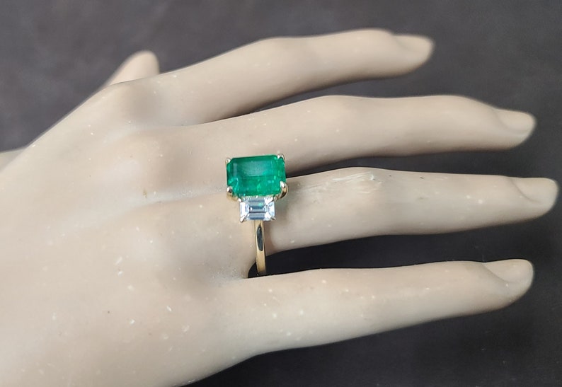 Op de afbeelding: Een gouden ring met een grote smaragdgroene edelsteen en twee kleinere, heldere edelstenen. De ring is op een hand van een mannequin.