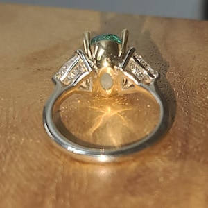May include: A close-up of a two-tone ring with a silver band and gold setting. The ring features a central green gemstone and smaller clear stones on either side. The ring is set against a warm, wooden surface.