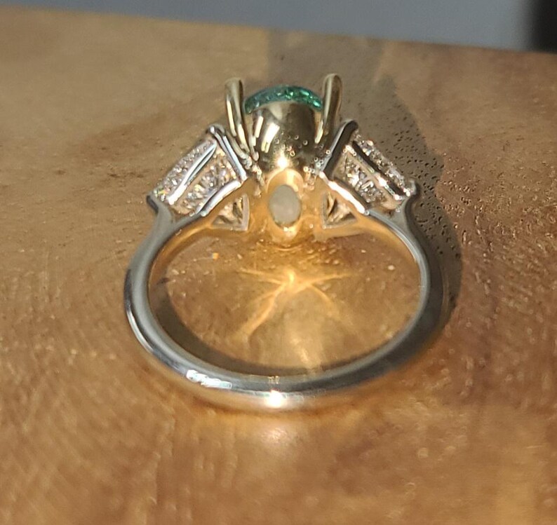May include: A close-up of a two-tone ring with a silver band and gold setting. The ring features a central green gemstone and smaller clear stones on either side. The ring is set against a warm, wooden surface.