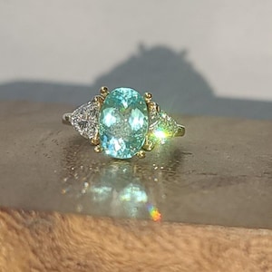 May include: A gold and silver ring featuring a large, oval-cut, light blue gemstone. Two smaller, triangular-shaped, diamond-studded accents flank the main stone. The ring is set on a wooden surface.
