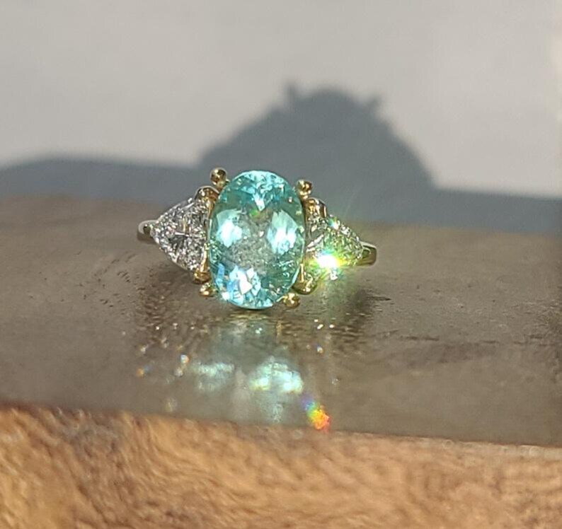 May include: A gold and silver ring featuring a large, oval-cut, light blue gemstone. Two smaller, triangular-shaped, diamond-studded accents flank the main stone. The ring is set on a wooden surface.