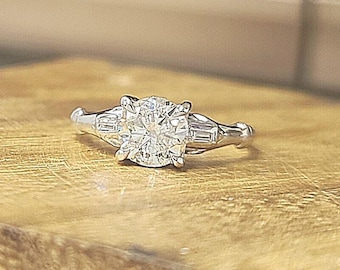 May include: A silver-toned ring featuring a large, round, faceted center stone held by four prongs. The band is accented on each side by two smaller, rectangular, baguette-cut stones set horizontally, creating a classic three-stone design.