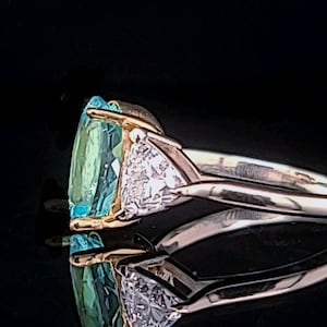 May include: A gold ring with a large, light blue gemstone and a smaller, triangular diamond. The ring is set against a black background, highlighting the sparkle of the gems. The ring's design is elegant and modern.