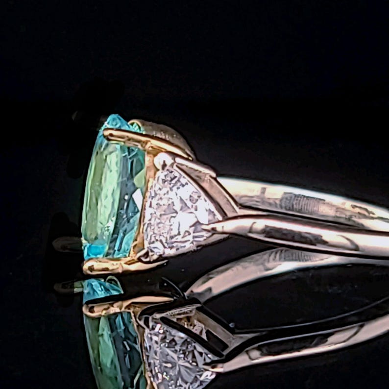 May include: A gold ring with a large, light blue gemstone and a smaller, triangular diamond. The ring is set against a black background, highlighting the sparkle of the gems. The ring's design is elegant and modern.