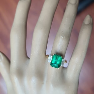 Op de afbeelding: Een gouden ring met een grote smaragdgroene edelsteen omringd door twee kleinere, heldere edelstenen.