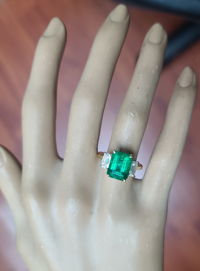 Op de afbeelding: Een gouden ring met een grote smaragdgroene edelsteen omringd door twee kleinere, heldere edelstenen.