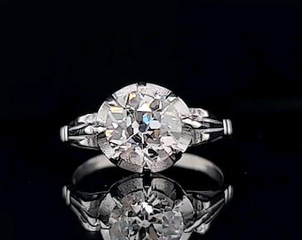May include: A platinum engagement ring with a large, round, brilliant-cut diamond set in a bezel setting. The diamond is surrounded by a halo of smaller diamonds.