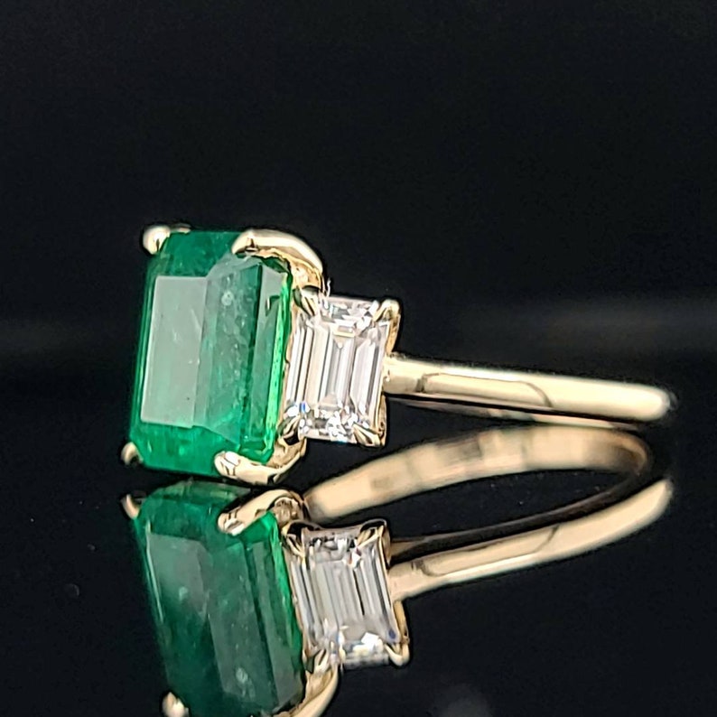 Op de afbeelding: Een gouden ring met een grote smaragdgroene edelsteen en twee kleinere, rechthoekige, heldere edelstenen.