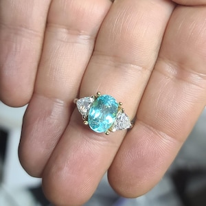 May include: A gold ring featuring a large, oval-cut, light blue gemstone flanked by two smaller, triangular, clear gemstones. The ring's setting has gold accents.