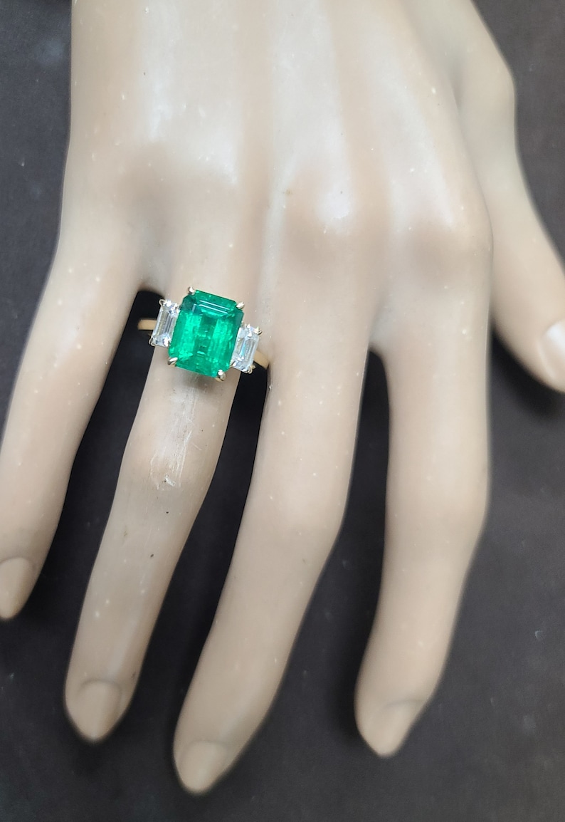 Op de afbeelding: Een gouden ring met een grote smaragdgroene edelsteen en twee kleinere baguette-geslepen diamanten aan weerszijden.