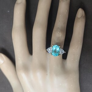 May include: A gold ring with a large, oval, light blue gemstone as the centerpiece. The ring has two smaller, triangular, white gemstones on either side of the main stone. The ring is set on a pale hand.