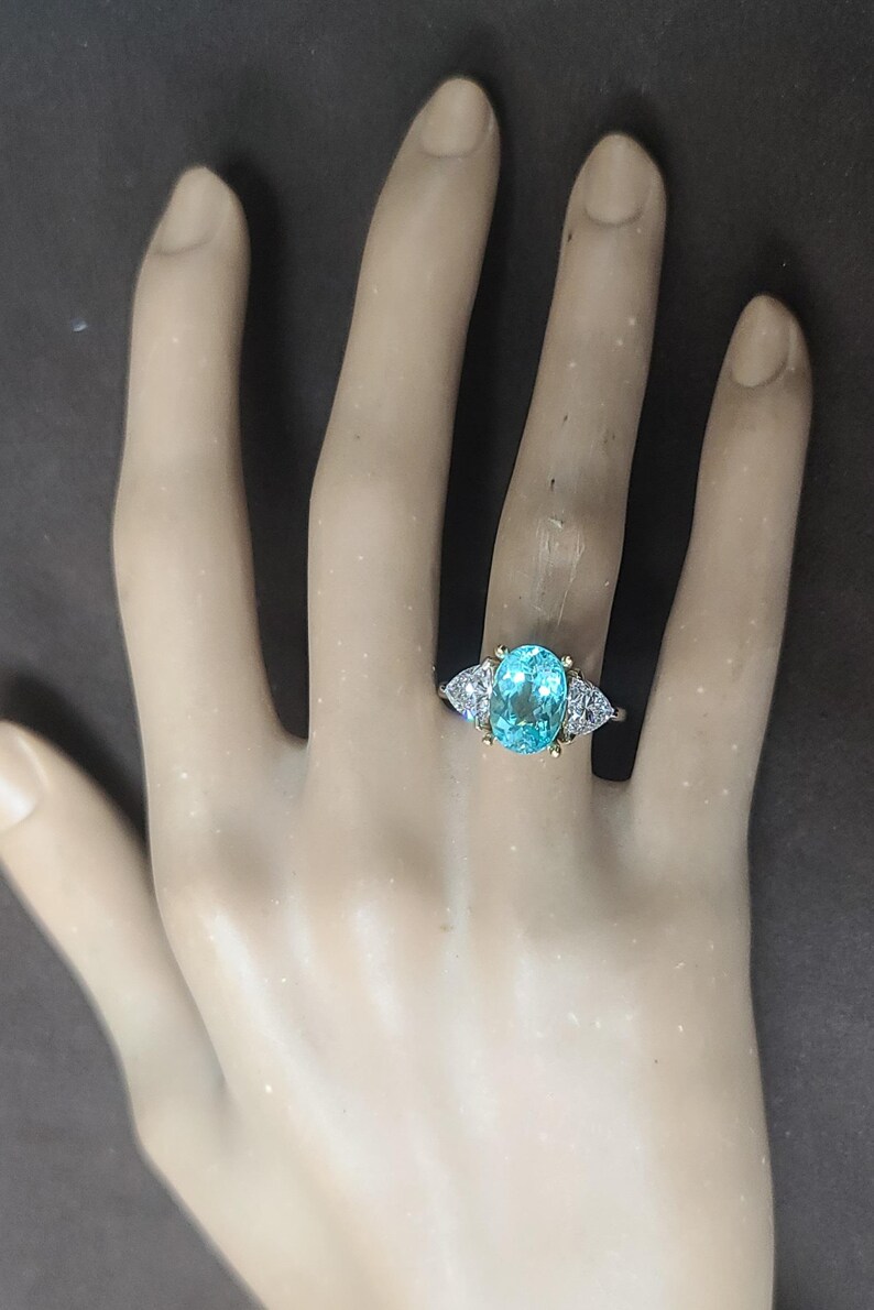 May include: A gold ring with a large, oval, light blue gemstone as the centerpiece. The ring has two smaller, triangular, white gemstones on either side of the main stone. The ring is set on a pale hand.