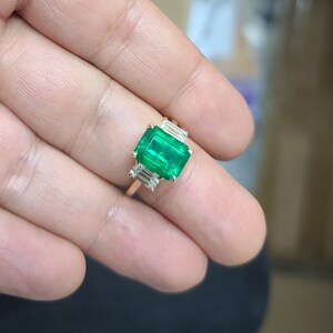 Op de afbeelding: Een gouden ring met een grote smaragdgroene edelsteen omringd door drie kleinere, rechthoekige, heldere edelstenen.