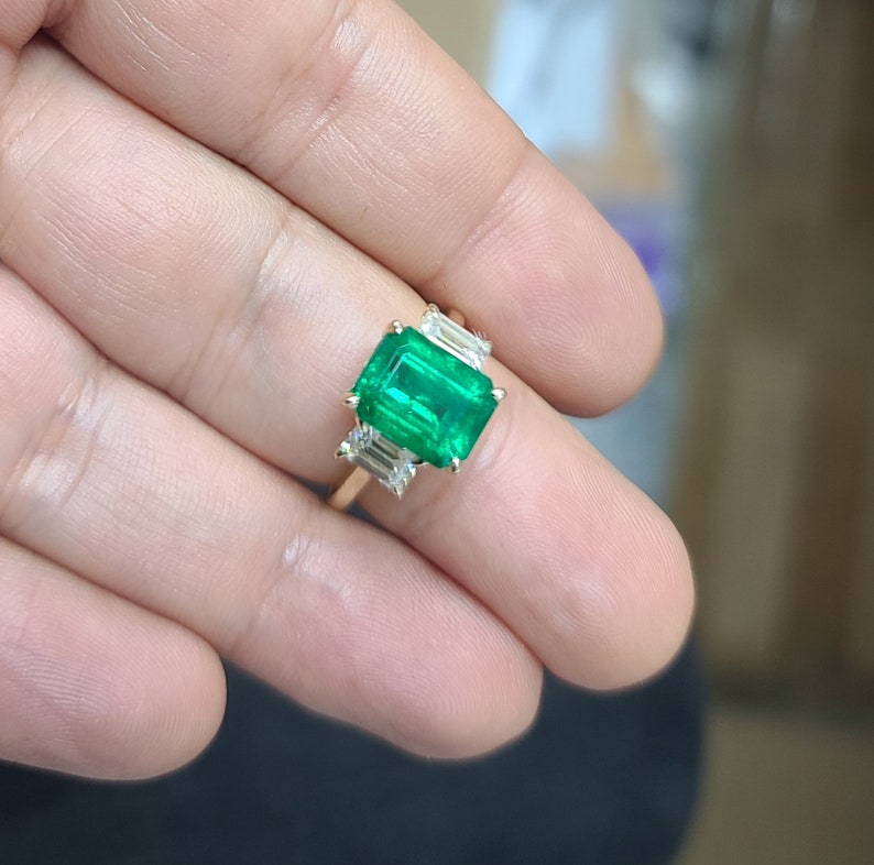 Op de afbeelding: Een gouden ring met een grote smaragdgroene edelsteen omringd door drie kleinere, rechthoekige, heldere edelstenen.