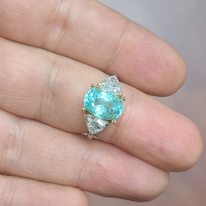May include: A silver ring featuring a large, oval, light blue gemstone as the centerpiece. The ring is accented with smaller, triangular, clear gemstones on either side of the main stone. The setting includes gold accents.