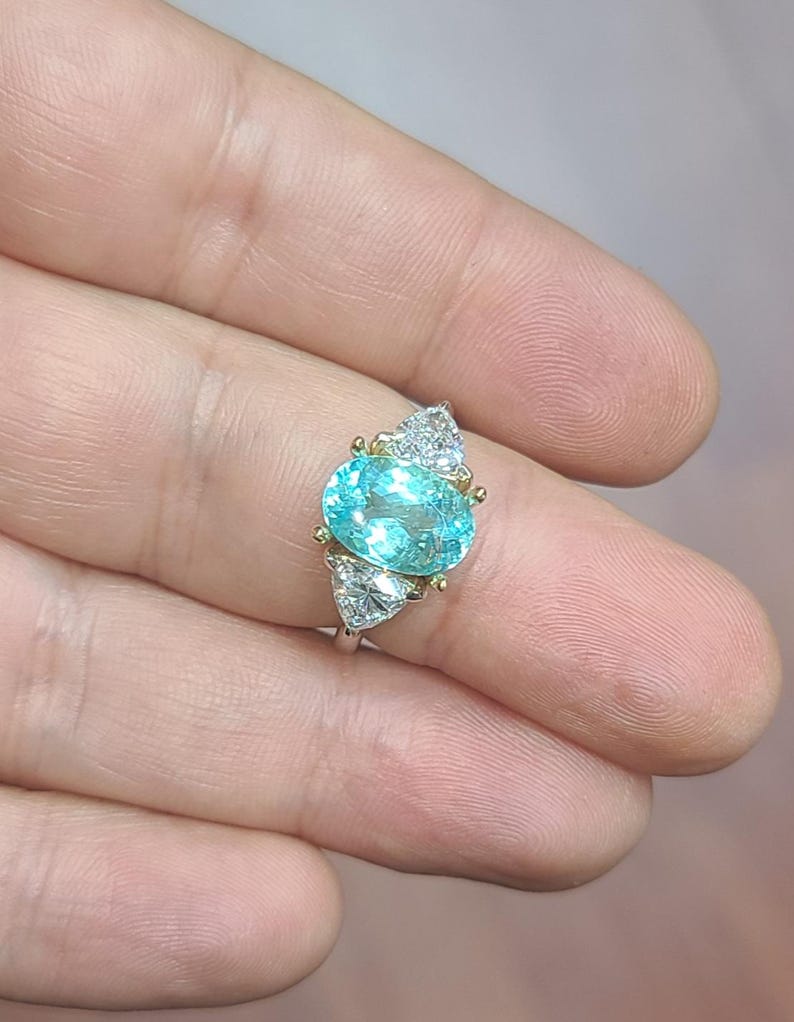 May include: A silver ring featuring a large, oval, light blue gemstone as the centerpiece. The ring is accented with smaller, triangular, clear gemstones on either side of the main stone. The setting includes gold accents.