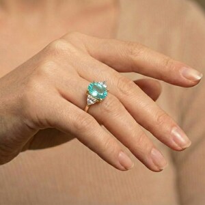 May include: A gold ring featuring a large, oval-cut, light blue gemstone as the centerpiece. The ring is accented with smaller, clear gemstones on either side of the main stone. The ring is set on a gold band.