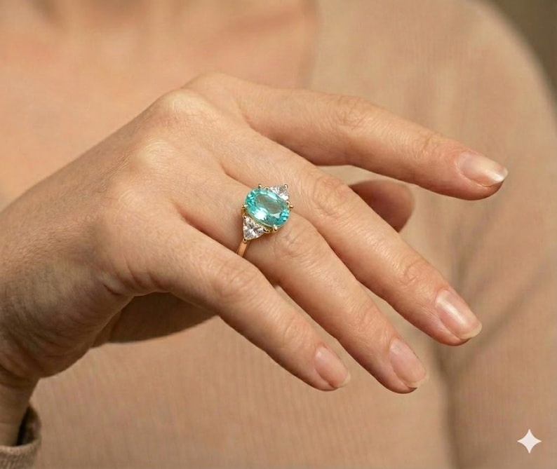 May include: A gold ring featuring a large, oval-cut, light blue gemstone as the centerpiece. The ring is accented with smaller, clear gemstones on either side of the main stone. The ring is set on a gold band.