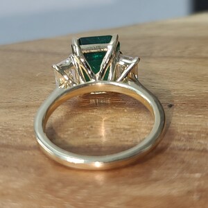 Op de afbeelding: Een gouden ring met een grote smaragd geslepen groene edelsteen, omringd door twee kleinere ronde diamanten.