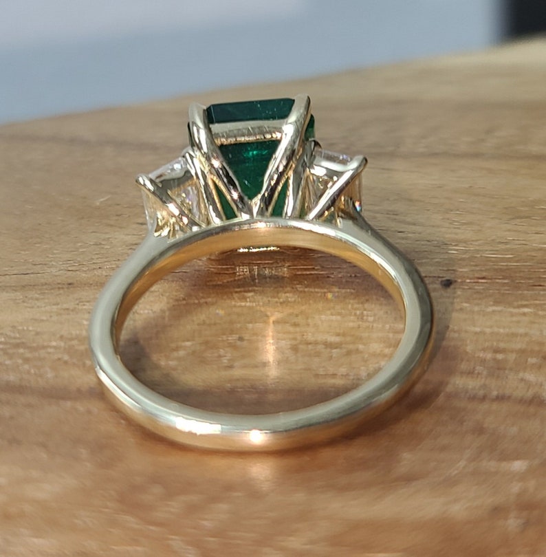 Op de afbeelding: Een gouden ring met een grote smaragd geslepen groene edelsteen, omringd door twee kleinere ronde diamanten.