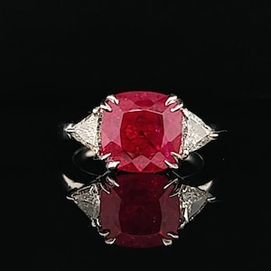 May include: A ring with a large, red, cushion-cut ruby surrounded by two smaller, triangular diamonds.