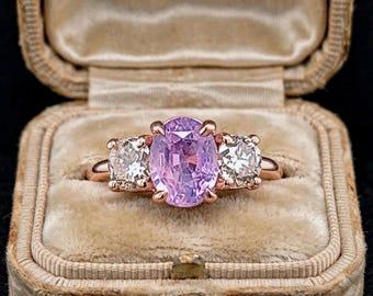 May include: A three-stone ring featuring a central oval-cut light purple gemstone flanked by two round-cut clear white stones. The stones are set in a polished rose gold band with four prongs securing the center stone and prongs for the side stones.
