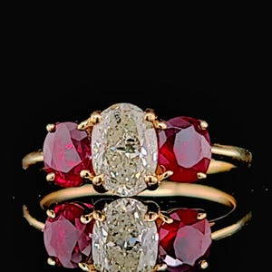 May include: A gold ring featuring a large, oval-cut diamond flanked by two round, red gemstones. The ring's design is classic, with the gemstones set in a prong setting. The ring is displayed against a black background.