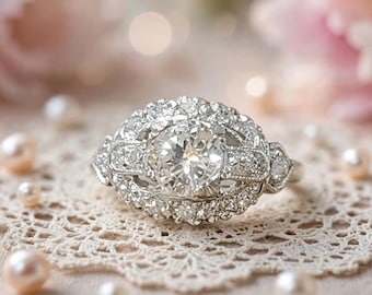 May include: A silver-toned ring featuring a large, round, faceted central stone surrounded by a halo of smaller, sparkling stones. The metal band is intricately detailed with milgrain edges and additional small stones along the shoulders.