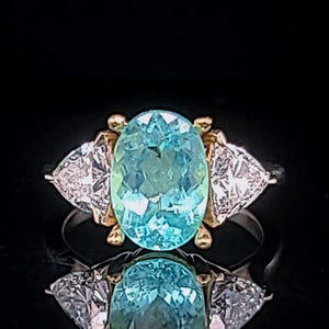 May include: A ring featuring a large, oval-cut, light blue gemstone as the centerpiece, flanked by two triangular, clear gemstones. The ring has a gold setting around the central stone and a silver band. The ring is displayed against a black background.