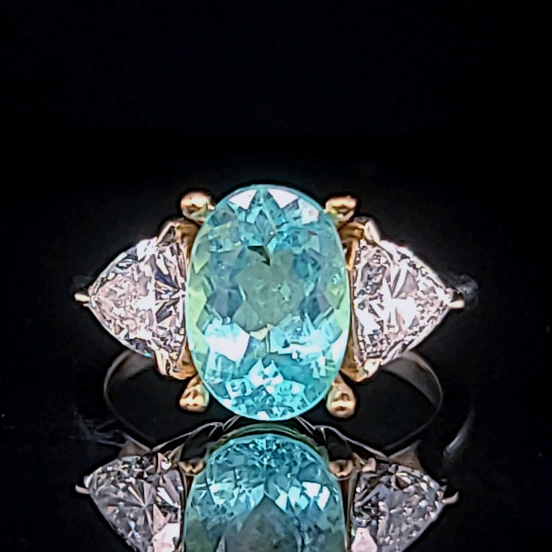 May include: A ring featuring a large, oval-cut, light blue gemstone as the centerpiece, flanked by two triangular, clear gemstones. The ring has a gold setting around the central stone and a silver band. The ring is displayed against a black background.